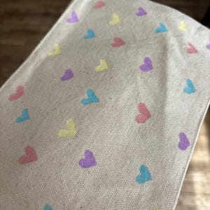 May include: A beige canvas tote bag with a repeating pattern of pastel pink, yellow, blue, and purple hearts.