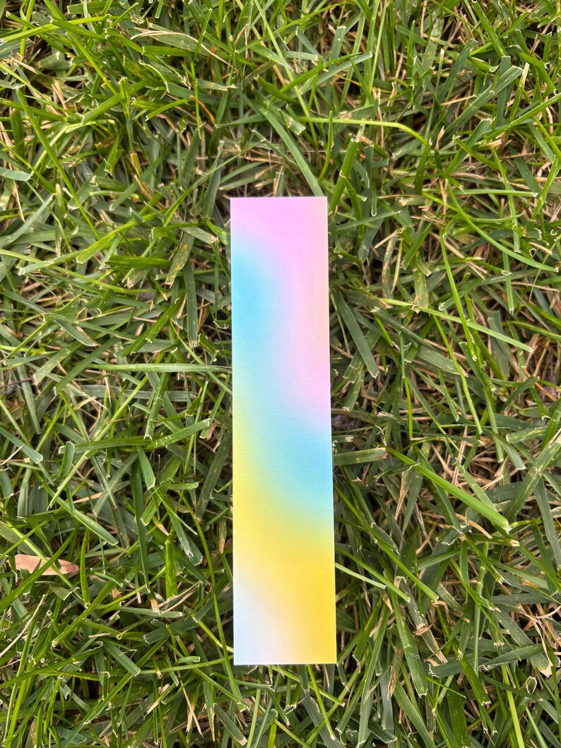 One gradient bookmark sitting on grass. Shows the different colors of the light blue, pink and yellow.