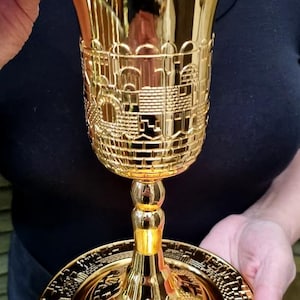 Pode incluir: Um cálice dourado com intrincados desenhos em relevo, segurado por uma pessoa. O cálice apresenta uma borda larga, um corpo detalhado com padrões arquitetônicos e uma base redonda. O cálice está sobre um prato a condizer.