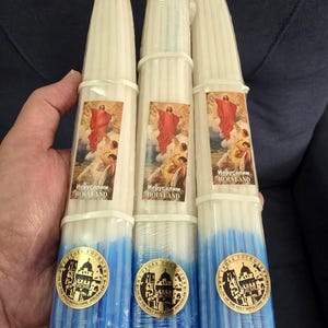 Könnte beinhalten: Drei Bündel weißer Stabkerzen mit blau getauchten Enden, in Klarsichtfolie verpackt. Jedes Bündel hat ein rechteckiges Etikett mit einer religiösen Szene und dem Text Holy Land sowie ein goldenes Siegel mit einem Kirchengebäude.