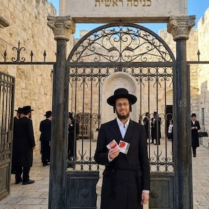 May include: A person wearing a black hat, white shirt, and long black coat stands before an ornate metal gate with a stone archway featuring Hebrew text. The person holds two small, square, red and white cards in their hands.