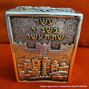 May include: A silver metal box with a textured surface and a gold inscription in Hebrew that reads "Shema Yisrael Adonai Eloheinu Adonai Echad". The box features a raised image of the Jerusalem skyline with a silver and gold finish.