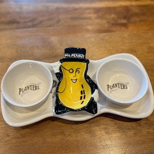 Peut inclure: Un plateau de service en céramique blanche avec deux petits bols et un plat en forme d'arachide. Les bols et le plat sont décorés du logo Planters et le plat en forme d'arachide présente un dessin animé de M. Peanut portant un chapeau haut de forme et des lunettes.