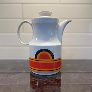 Peut inclure: Cafetière en céramique blanche avec un motif géométrique orange, jaune et noir. Le motif présente une forme d'arche arrondie avec un cercle plus petit à l'intérieur.