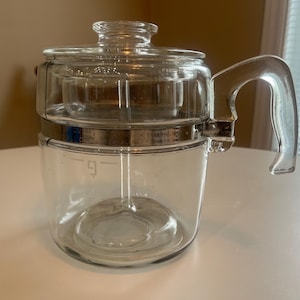 May include: A clear glass coffee percolator with a glass handle and lid. The percolator has a metal band and a measurement indicator for up to 9 cups. The design is vintage and functional.