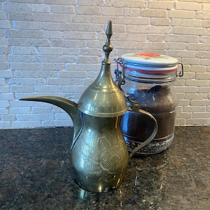 May include: A brass Arabic coffee pot with an ornate design and a lid. The pot is sitting on a dark countertop next to a glass jar with a red and white lid.