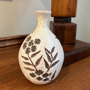 May include: A small, off-white ceramic vase with a narrow neck and rounded body. The vase is decorated with brown floral and leaf patterns. The vase is approximately 15 cm tall.