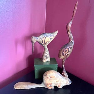 May include: Three wooden animal sculptures: a bird with a long neck, a bird with a short neck and a beaver. The sculptures are made of wood with a natural finish and are displayed on a black surface.