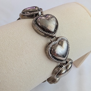 May include: A silver-toned link bracelet featuring multiple puffed heart-shaped charms. Each heart is framed by a twisted rope-style border and connected by small metal rings, creating a continuous chain of polished, reflective heart elements.