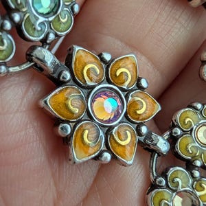 May include: A silver-toned metal bracelet featuring a series of linked flower-shaped charms. Each charm is decorated with yellow and orange enamel petals and a central iridescent crystal. The links are connected by small silver-toned metal loops.