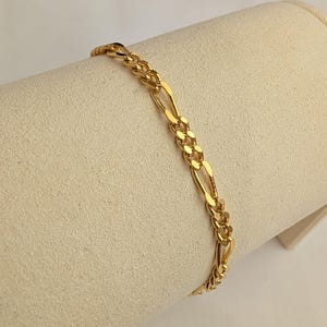May include: A gold-toned Figaro chain bracelet featuring a repeating pattern of three small circular links followed by one elongated oval link. The metal has a polished, reflective finish that catches the light, creating a bright, metallic appearance.