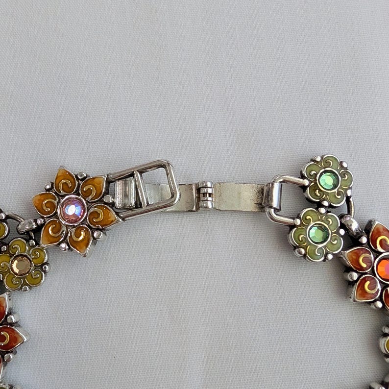 May include: A silver-toned metal bracelet featuring a series of connected flower-shaped links. Each flower is filled with colorful enamel in shades of orange, yellow, and green, and accented with small, round, sparkling crystals in the center.