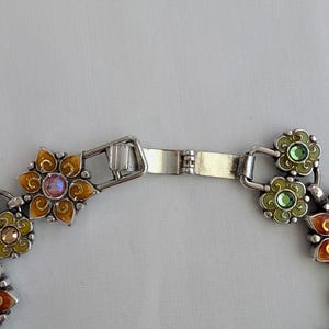 May include: A silver-toned metal bracelet featuring a series of connected flower-shaped links. Each link is filled with colorful enamel in shades of orange, yellow, and green, and centered with a small, sparkling round crystal in matching tones.