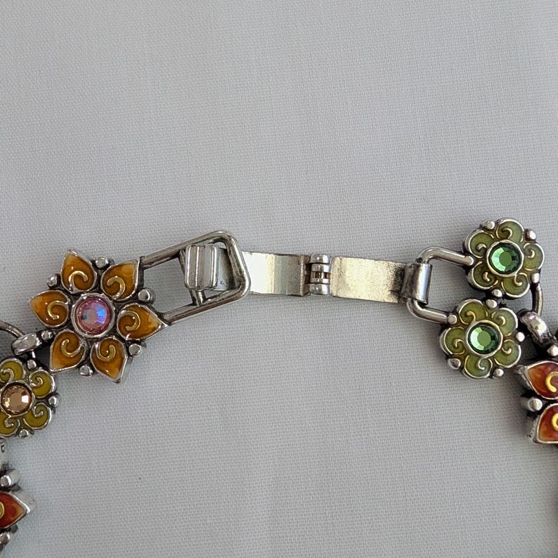 May include: A silver-toned metal bracelet featuring a series of connected flower-shaped links. Each link is filled with colorful enamel in shades of orange, yellow, and green, and centered with a small, sparkling round crystal in matching tones.