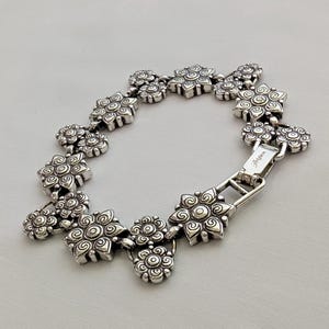 May include: A silver-toned metal link bracelet featuring a series of repeating floral-shaped charms with embossed spiral and circular patterns. The bracelet includes a rectangular clasp mechanism with the brand name Brighton engraved on the surface.