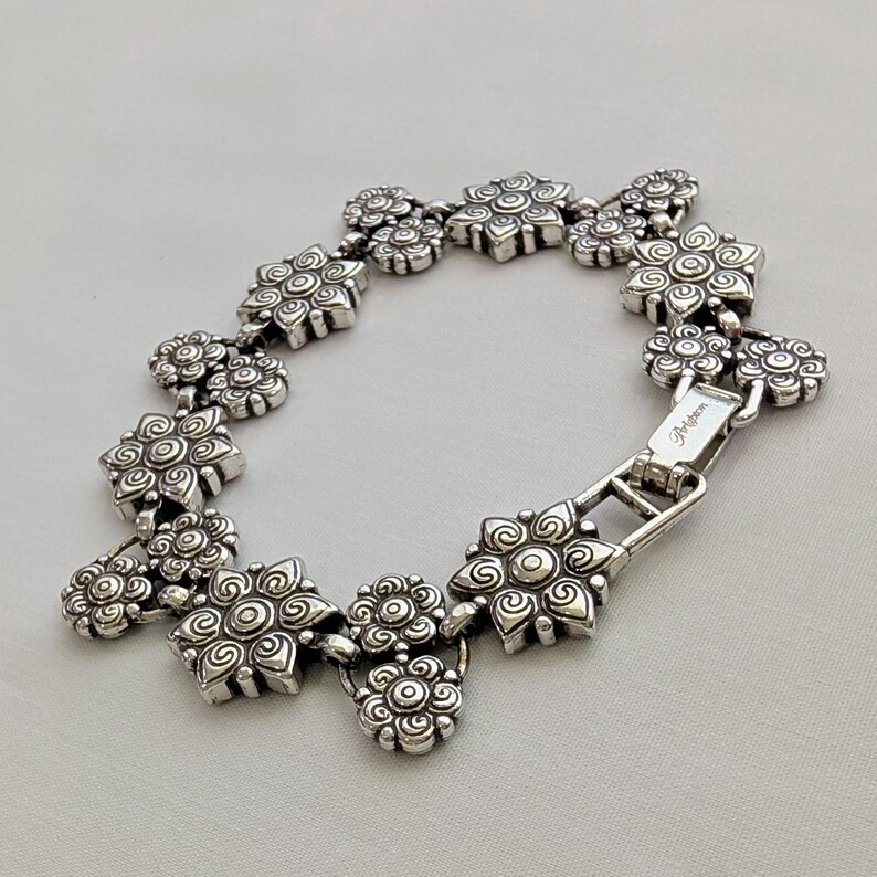 May include: A silver-toned metal link bracelet featuring a series of repeating floral-shaped charms with embossed spiral and circular patterns. The bracelet includes a rectangular clasp mechanism with the brand name Brighton engraved on the surface.