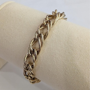 May include: A gold-colored chain bracelet with a thick, interwoven design. The bracelet is displayed on a cream-colored surface, showcasing its polished, shiny appearance. The links are oval-shaped and tightly connected, creating a bold, statement piece of jewelry.