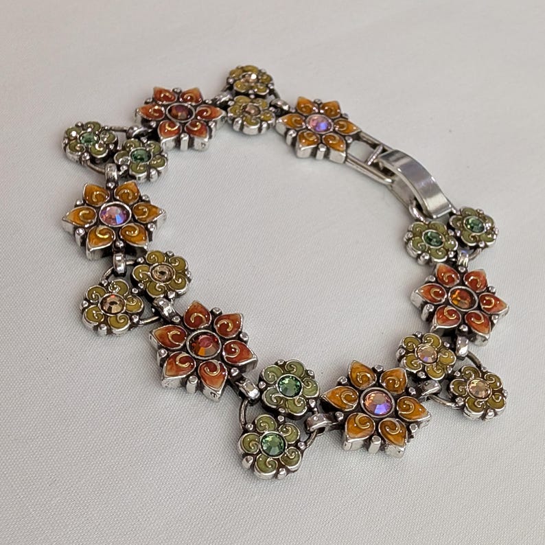 May include: A silver-tone link bracelet featuring a series of connected flower-shaped charms. Each flower is filled with orange, yellow, or green enamel and accented with a central round crystal. The bracelet includes a secure metal fold-over clasp.