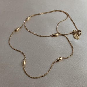 May include: A delicate gold-colored necklace featuring a fine chain with evenly spaced, oval-shaped beads. The necklace has a simple design with a small tag near the clasp. The jewelry is displayed on a light-colored surface.