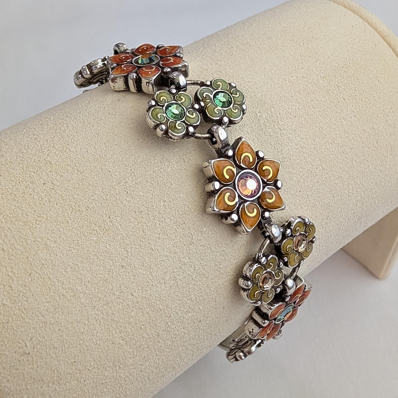 May include: A silver-toned link bracelet featuring alternating flower-shaped charms. The charms are decorated with orange and light green enamel and set with small, round, faceted crystals in the center of each floral design.
