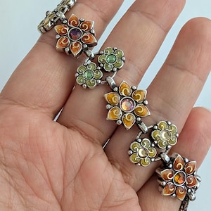 May include: A silver-toned link bracelet featuring alternating flower-shaped charms. The charms are decorated with orange and green enamel and set with central iridescent crystals. The metal links connect each floral element in a repeating pattern.