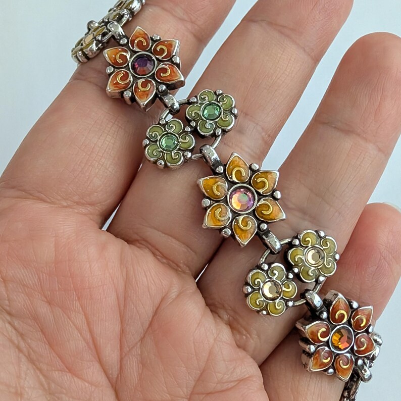 May include: A silver-toned link bracelet featuring alternating flower-shaped charms. The charms are decorated with orange and green enamel and set with central iridescent crystals. The metal links connect each floral element in a repeating pattern.