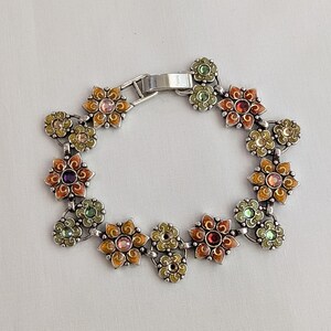 May include: A silver-toned metal link bracelet featuring a series of interconnected flower-shaped charms. Each flower is decorated with colorful enamel in shades of orange, yellow, and green, and centered with a small, round, multi-colored gemstone.