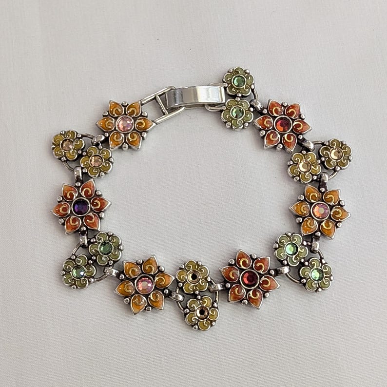 May include: A silver-toned metal link bracelet featuring a series of interconnected flower-shaped charms. Each flower is decorated with colorful enamel in shades of orange, yellow, and green, and centered with a small, round, multi-colored gemstone.