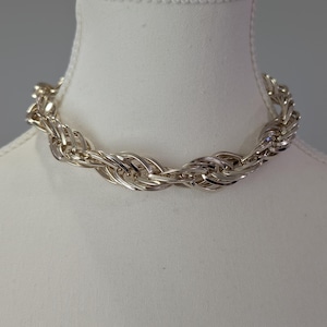 May include: A silver-toned necklace featuring a thick, braided rope chain design. The metallic links are intricately woven together to create a continuous, textured circular band that rests evenly around the neck area of the display form.
