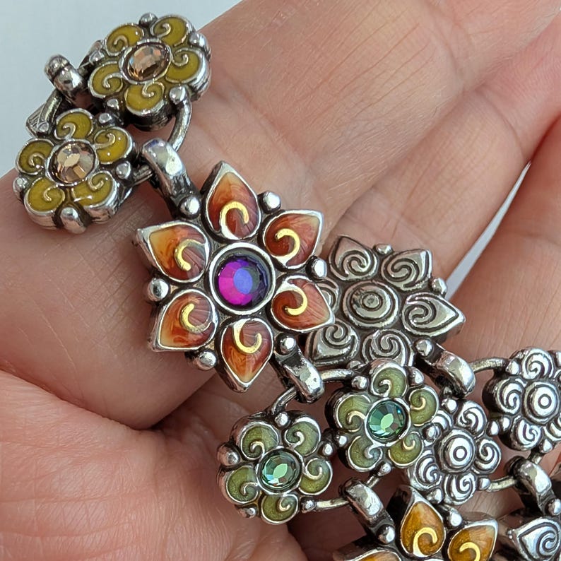 May include: A silver-toned link bracelet featuring a series of floral-shaped charms. The charms are decorated with yellow, orange, and green enamel, accented with small, round, multi-colored crystals in shades of purple, green, and amber.