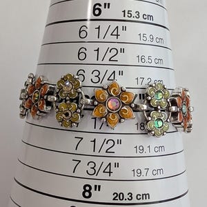 May include: A silver-tone link bracelet featuring alternating flower-shaped charms in orange and light green enamel with central iridescent crystals. The bracelet is positioned on a sizing cone, indicating a length of approximately 7 inches.