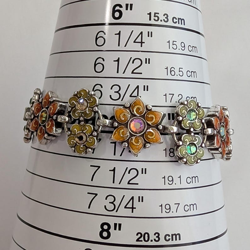 May include: A silver-tone link bracelet featuring alternating flower-shaped charms in orange and light green enamel with central iridescent crystals. The bracelet is positioned on a sizing cone, indicating a length of approximately 7 inches.