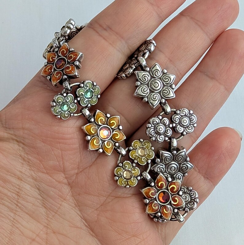 May include: A silver-toned link bracelet featuring a series of interconnected flower-shaped charms. The charms include solid silver floral designs and colorful enamel-filled petals in shades of orange, yellow, and green, with small embedded crystals.