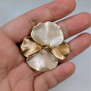May include: A gold and cream-colored flower-shaped brooch. The brooch has four petals, with two petals in a gold color and two petals in a cream color. The center of the brooch has three small gold beads.