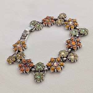 May include: A silver-toned link bracelet featuring a series of connected flower-shaped charms. The flowers are decorated with orange, yellow, and green enamel accents and centered with small, iridescent, multi-colored round crystals.
