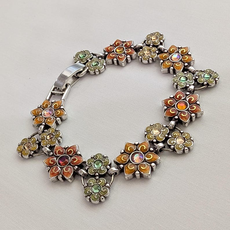 May include: A silver-toned link bracelet featuring a series of connected flower-shaped charms. The flowers are decorated with orange, yellow, and green enamel accents and centered with small, iridescent, multi-colored round crystals.