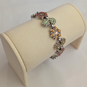 May include: A silver-toned link bracelet featuring a series of alternating flower-shaped charms. Each flower is decorated with colorful enamel in shades of orange and green, with small sparkling crystals set in the center of each individual blossom.