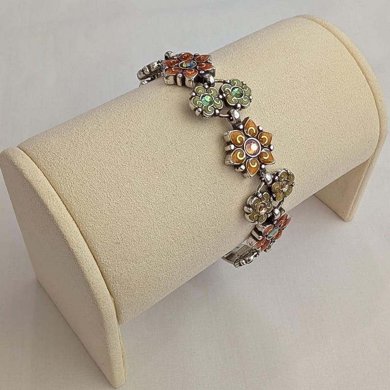 May include: A silver-toned link bracelet featuring a series of alternating flower-shaped charms. Each flower is decorated with colorful enamel in shades of orange and green, with small sparkling crystals set in the center of each individual blossom.