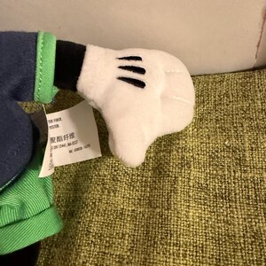 May include: A plush toy hand in white with three black lines on the palm. The hand is part of a larger stuffed character, with green and blue fabric visible. A tag with text is attached.