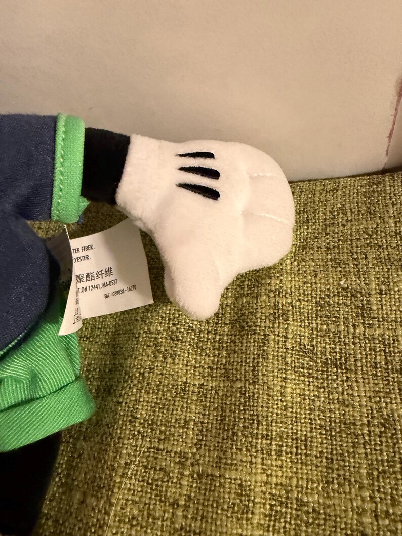 May include: A plush toy hand in white with three black lines on the palm. The hand is part of a larger stuffed character, with green and blue fabric visible. A tag with text is attached.