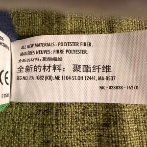 May include: A white tag with black text, including "ALL NEW MATERIALS: POLYESTER FIBER" and "REG NO. PA 1082 (KR): ME 1104-ST.OH 12441, MA-0537". The tag is on a green textured fabric. The tag also includes text in French and Chinese.