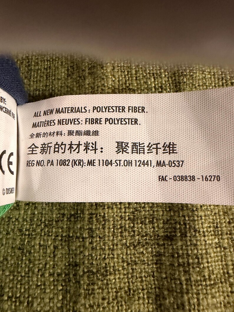May include: A white tag with black text, including "ALL NEW MATERIALS: POLYESTER FIBER" and "REG NO. PA 1082 (KR): ME 1104-ST.OH 12441, MA-0537". The tag is on a green textured fabric. The tag also includes text in French and Chinese.