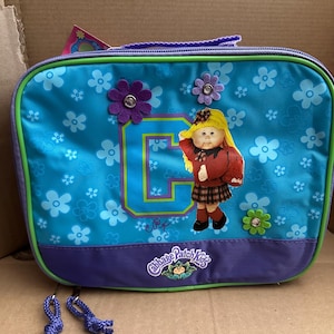 May include: A blue lunch bag with a Cabbage Patch Kids logo and a flower pattern. The bag features a large letter C with a Cabbage Patch Kid doll wearing a red dress and a yellow hat.