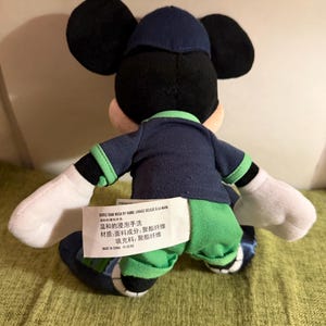 May include: A plush Mickey Mouse doll, viewed from the back, wearing a navy blue outfit with green accents. The doll has black ears, a navy blue hat, and white gloves. A tag with text in multiple languages is attached to the doll.