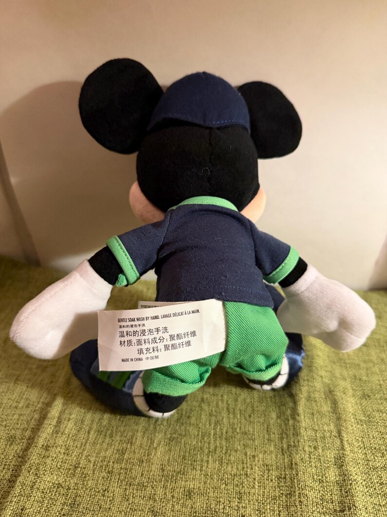 May include: A plush Mickey Mouse doll, viewed from the back, wearing a navy blue outfit with green accents. The doll has black ears, a navy blue hat, and white gloves. A tag with text in multiple languages is attached to the doll.