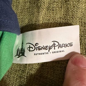 May include: A white tag with the text "Disney Parks AUTHENTIC &bull; ORIGINAL" in black. The tag has a green castle logo on the left side. The tag is attached to a green and blue fabric.