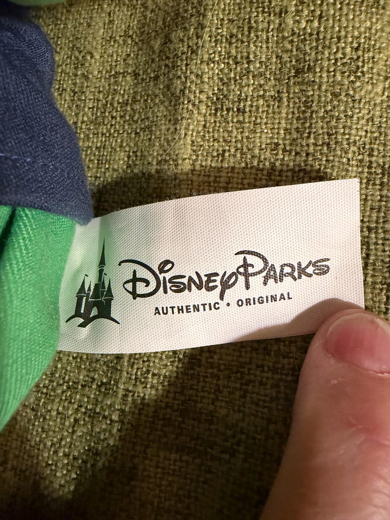 May include: A white tag with the text "Disney Parks AUTHENTIC &bull; ORIGINAL" in black. The tag has a green castle logo on the left side. The tag is attached to a green and blue fabric.