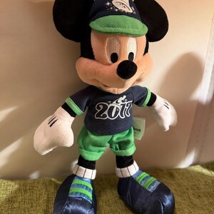 May include: A Mickey Mouse plush toy wearing a navy blue baseball cap and matching jersey with the number 2017. The plush has green shorts and shoes with blue and green stripes. The iconic character has black ears and white gloves.
