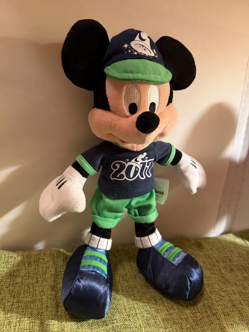 May include: A Mickey Mouse plush toy wearing a navy blue baseball cap and matching jersey with the number 2017. The plush has green shorts and shoes with blue and green stripes. The iconic character has black ears and white gloves.