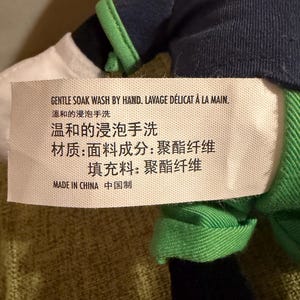 May include: A plush toy with a tag that reads "GENTLE SOAK WASH BY HAND" in English and French. The tag also includes Chinese characters. The toy has a navy blue and green outfit with white and black accents.